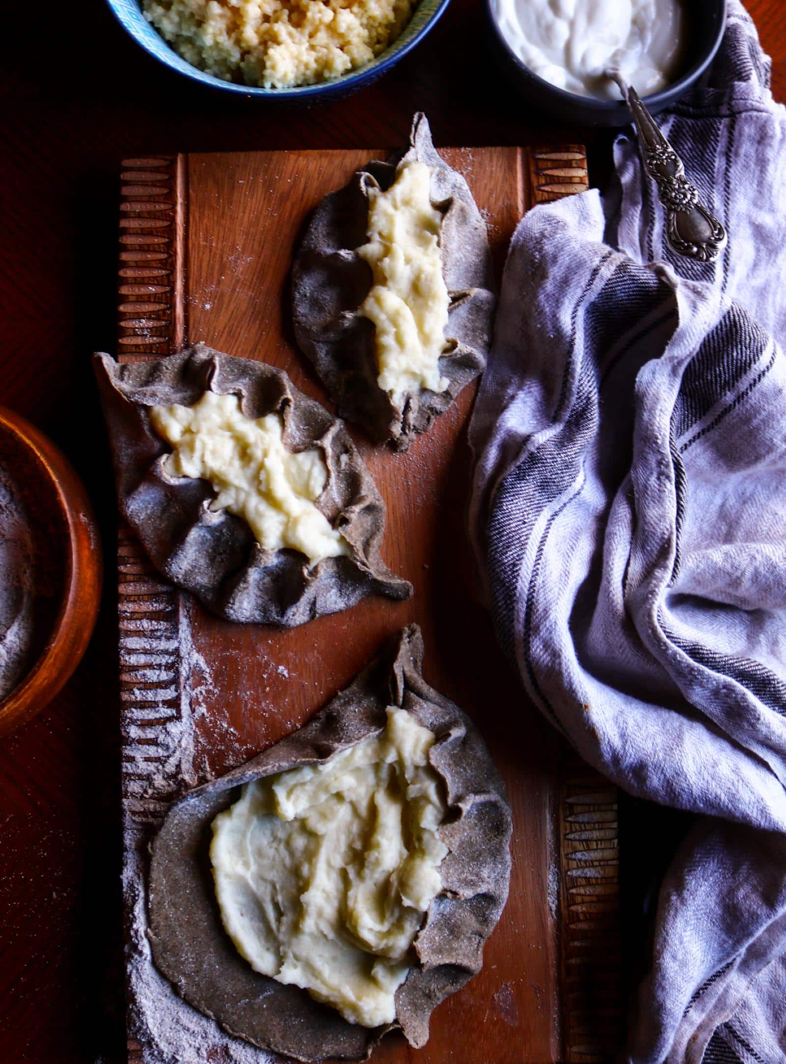 Rustic Karelian rye potato pies being shaped on a wooden board, with hand-pinched rye dough and creamy mashed potato filling, surrounded by flour, bowls of filling, and a linen kitchen towel.