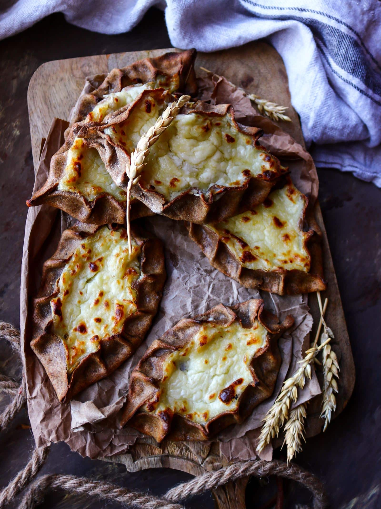 Rustic Karelian rye potato pies with hand-pinched rye crusts and creamy mashed potato filling, baked until golden and arranged on parchment on a wooden board.