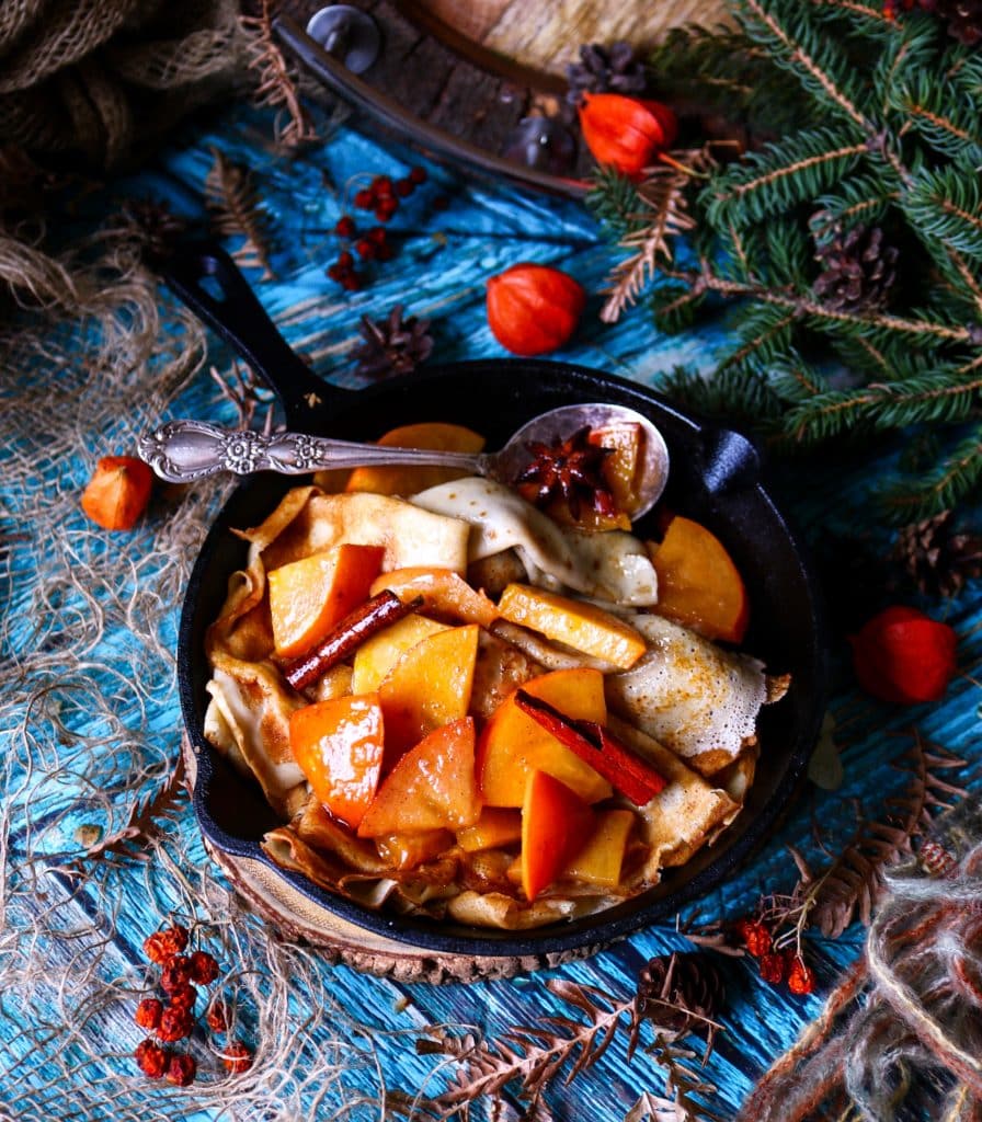 Honey Persimmon Crêpes in a cast-iron skillet topped with warm honey persimmon sauce, cinnamon sticks, and star anise, styled with rustic winter décor and evergreen branches.
