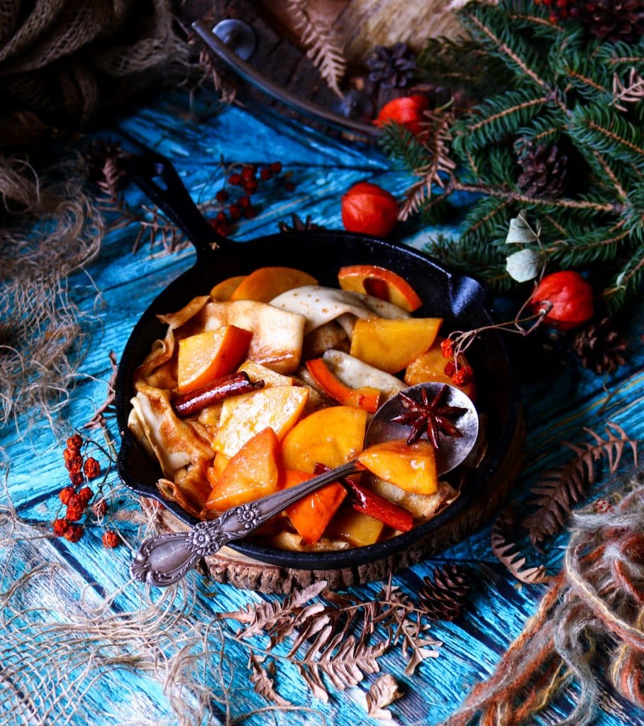 Honey Persimmon Crêpes in a cast-iron skillet topped with warm honey persimmon sauce, cinnamon sticks, and star anise, styled with rustic winter décor and evergreen branches.