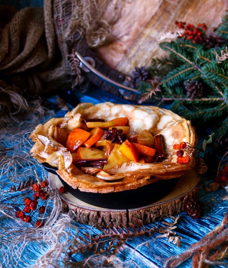 Honey Persimmon Crêpes in a cast-iron skillet topped with warm honey persimmon sauce, cinnamon sticks, and star anise, styled with rustic winter décor and evergreen branches.