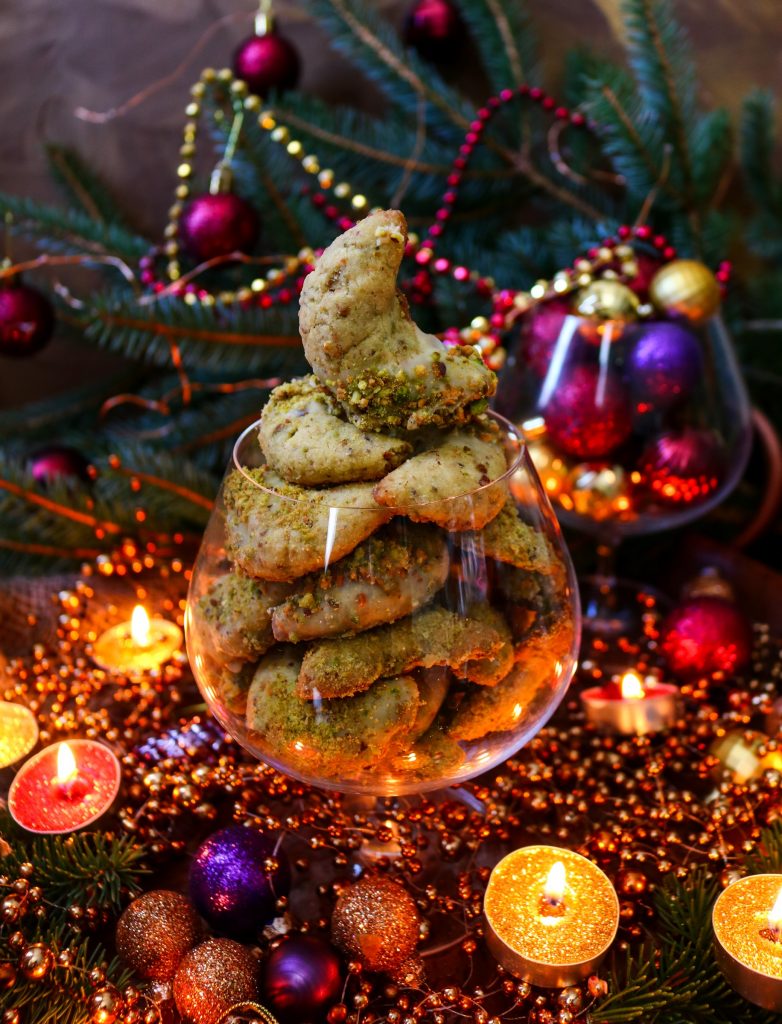 A glass filled with Festive Pistachio Crescent Cookies coated in crushed pistachios, surrounded by glowing candles, Christmas ornaments, and twinkling holiday lights for a warm festive atmosphere.