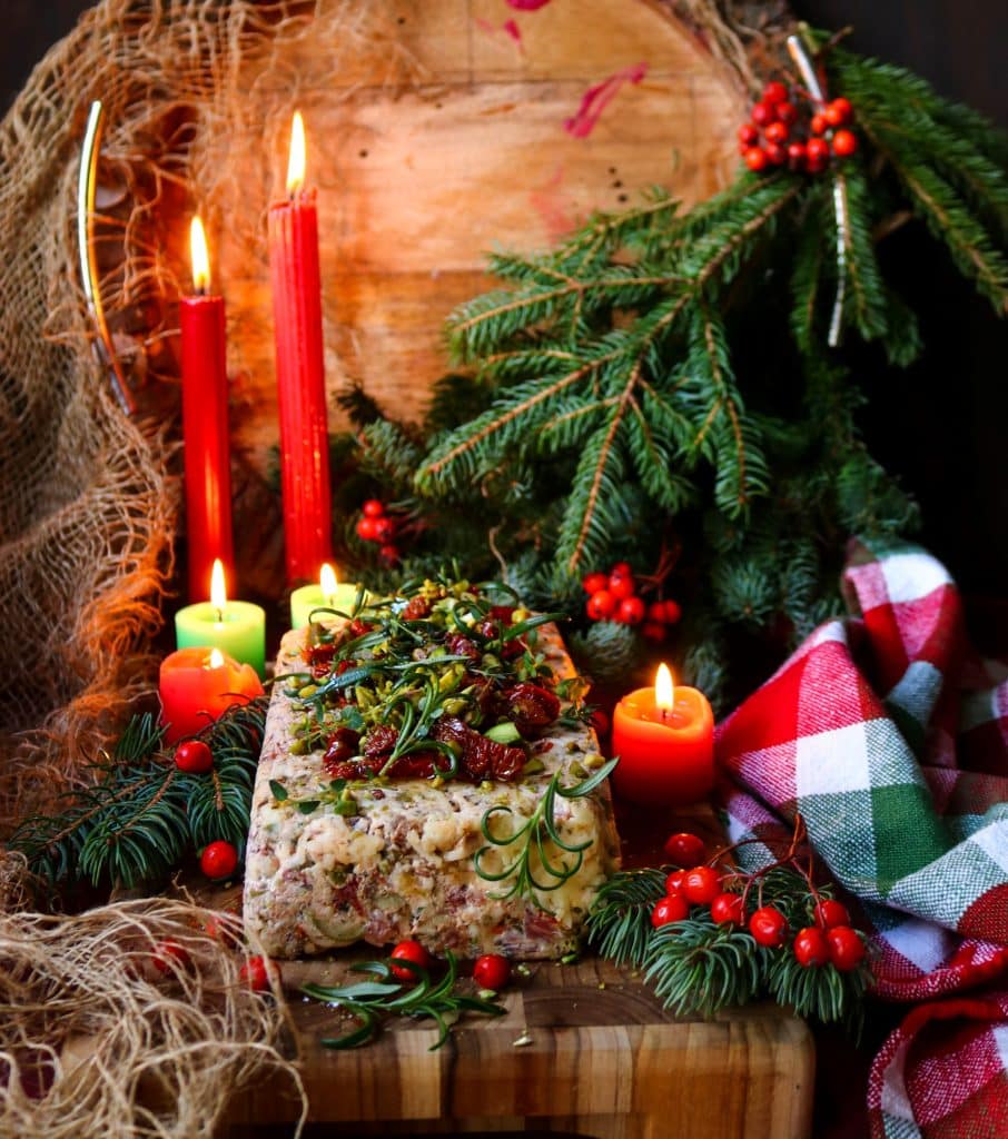 Festive Italian cheese log topped with pistachios, sun-dried tomatoes, and rosemary, surrounded by glowing red candles and evergreen branches.