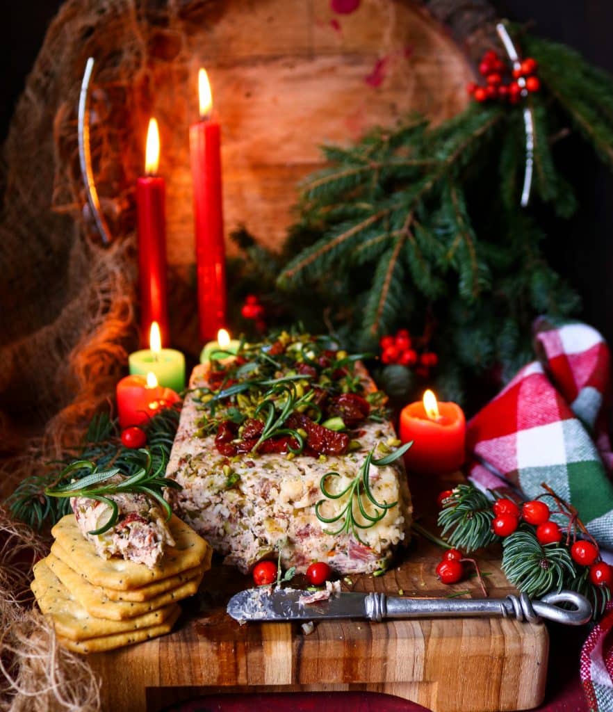 Elegant festive Italian cheese log served with crackers, topped with pistachios, tomatoes, and rosemary amid glowing candles and Christmas decor