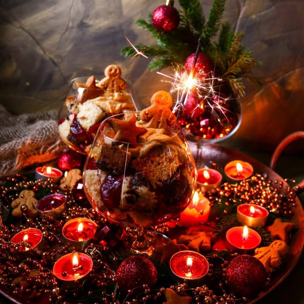 Holiday dessert trifles in glasses decorated with gingerbread cookies and sparklers, set on a tray with glowing candles, ornaments, and festive greenery.