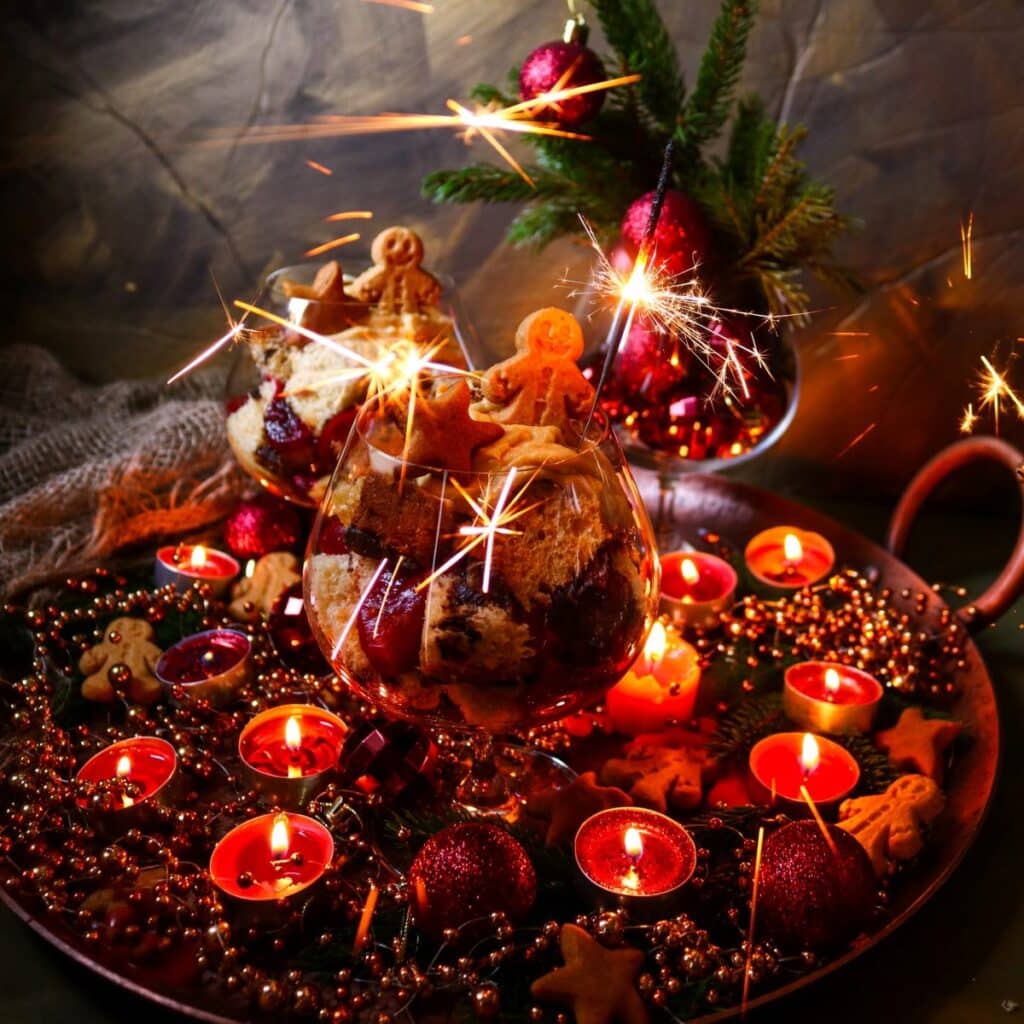 Holiday dessert trifles in glasses decorated with gingerbread cookies and sparklers, set on a tray with glowing candles, ornaments, and festive greenery.