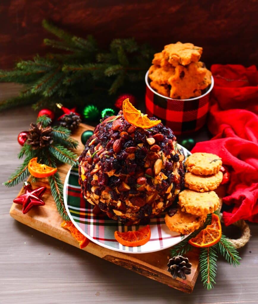 A festive cranberry almond cheese ball coated in chopped almonds and dried cranberries, garnished with a slice of orange and surrounded by holiday cookies, candles, and evergreens — a bright and elegant Christmas appetizer.