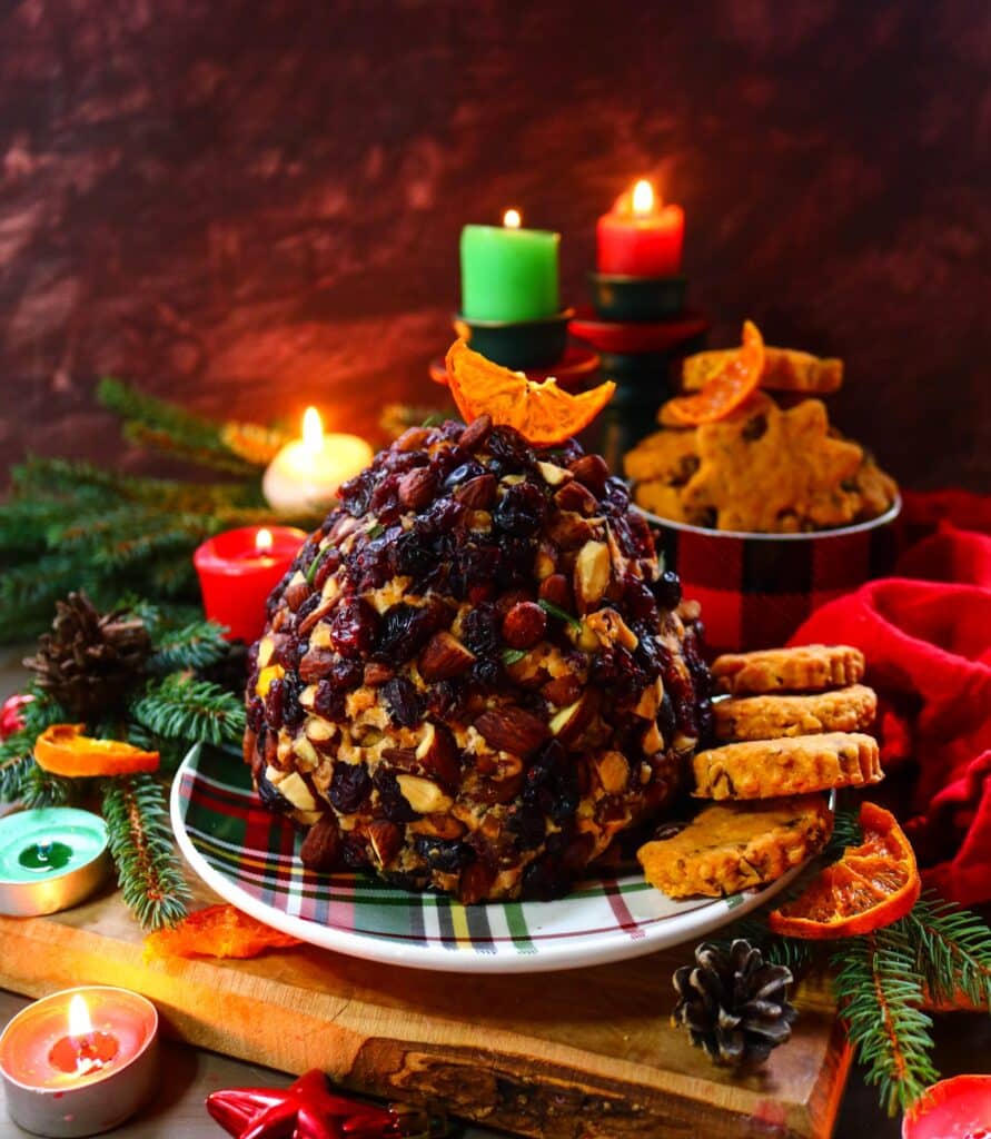 A festive cranberry almond cheese ball coated in chopped almonds and dried cranberries, garnished with a slice of orange and surrounded by holiday cookies, candles, and evergreens — a bright and elegant Christmas appetizer.