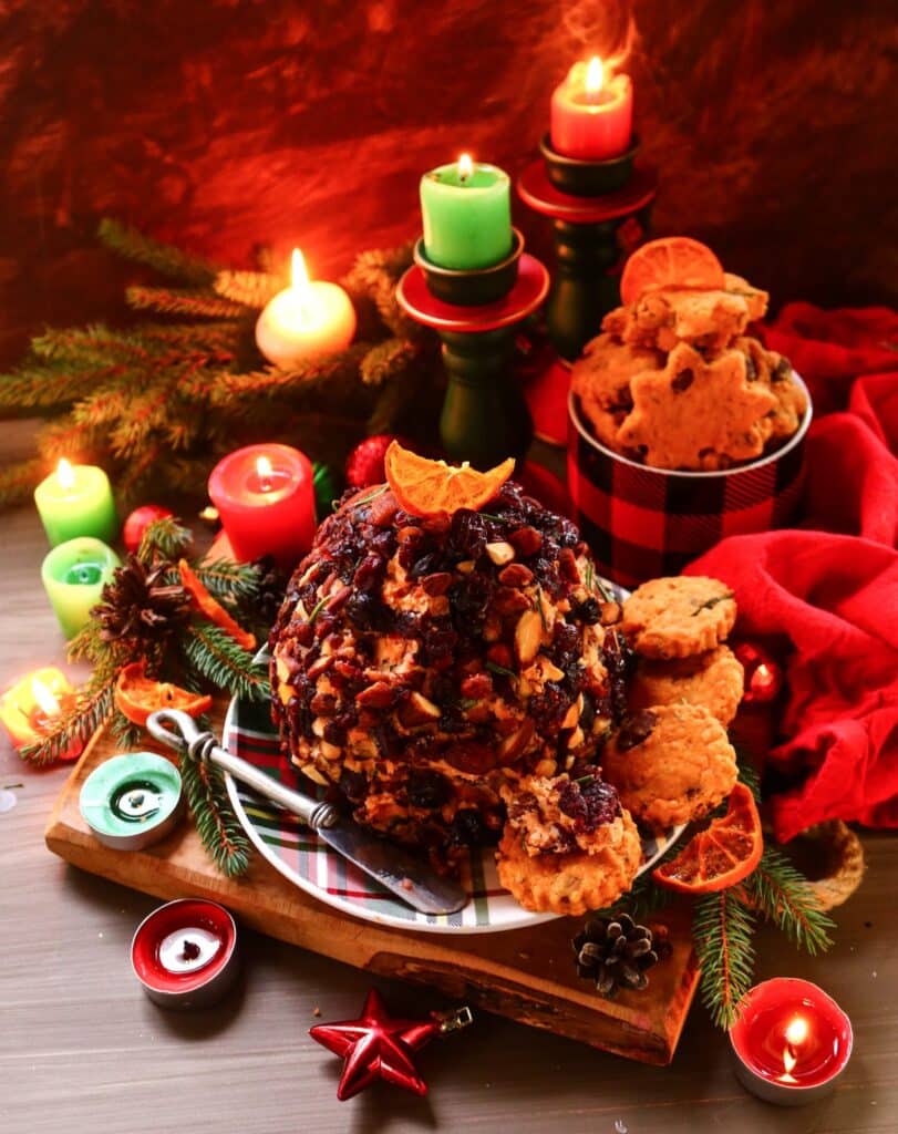 A festive cranberry almond cheese ball coated in chopped almonds and dried cranberries, garnished with a slice of orange and surrounded by holiday cookies, candles, and evergreens — a bright and elegant Christmas appetizer.