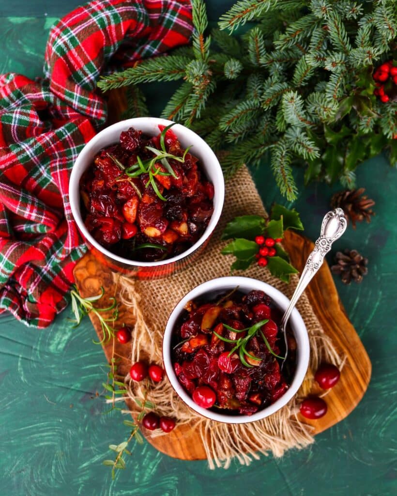 Overhead view of Festive Cranberry Almond Relish with caramelized onions, rosemary, and honey in plaid bowls on a wooden board with holiday greenery, cranberries, and tartan fabric — a vibrant red Christmas relish perfect for festive entertaining.