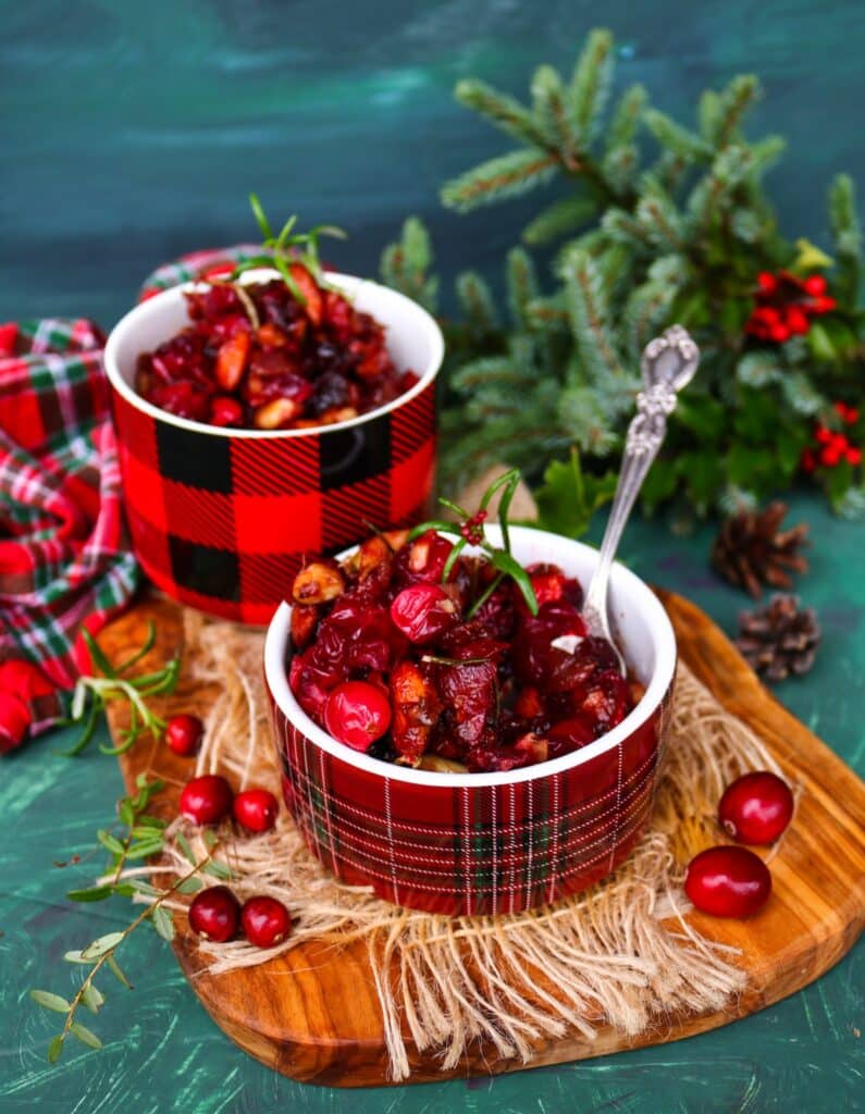 Festive Cranberry Almond Relish with caramelized onions, rosemary, and honey served in plaid holiday bowls on a wooden board with fresh cranberries and pine branches — a vibrant red Christmas condiment perfect for holiday entertaining.