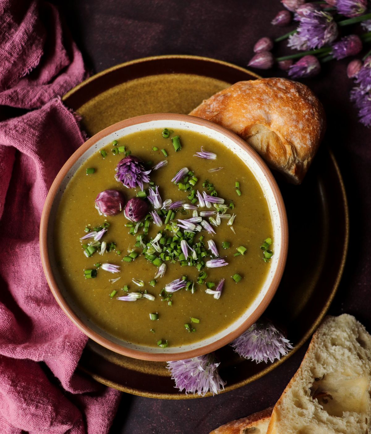 Creamy potato chive soup garnished with fresh chives and chive blossoms, served with crusty bread on a dark background.