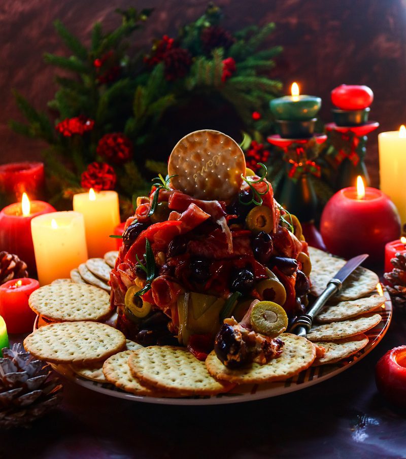 A festive antipasto cheese ball decorated with olives, prosciutto, and roasted peppers, served with crackers and surrounded by glowing candles and holiday greenery for a cozy Christmas presentation.