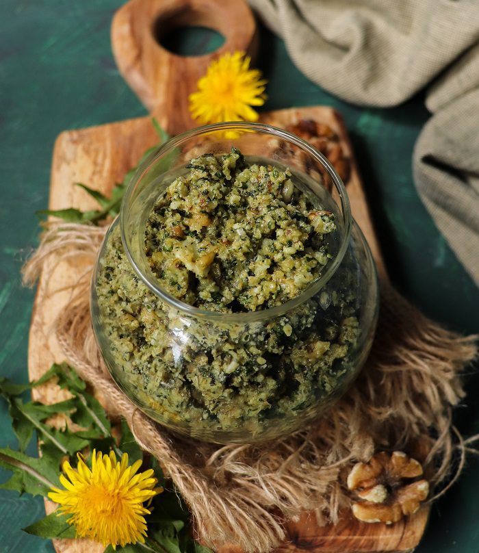 Dandelion greens pesto with walnuts in a glass jar, topped with a fresh yellow dandelion flower.