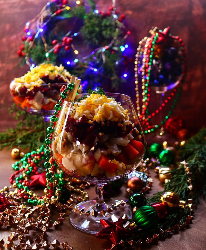 Layered herring salad with beets, potatoes, carrots and eggs served in elegant glasses, styled with festive holiday decorations and colourful Christmas lights.