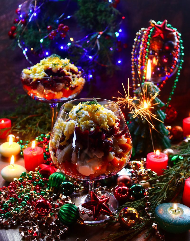 Layered herring salad with beets, potatoes, carrots and eggs served in elegant glasses, styled with festive holiday decorations and colourful Christmas lights.
