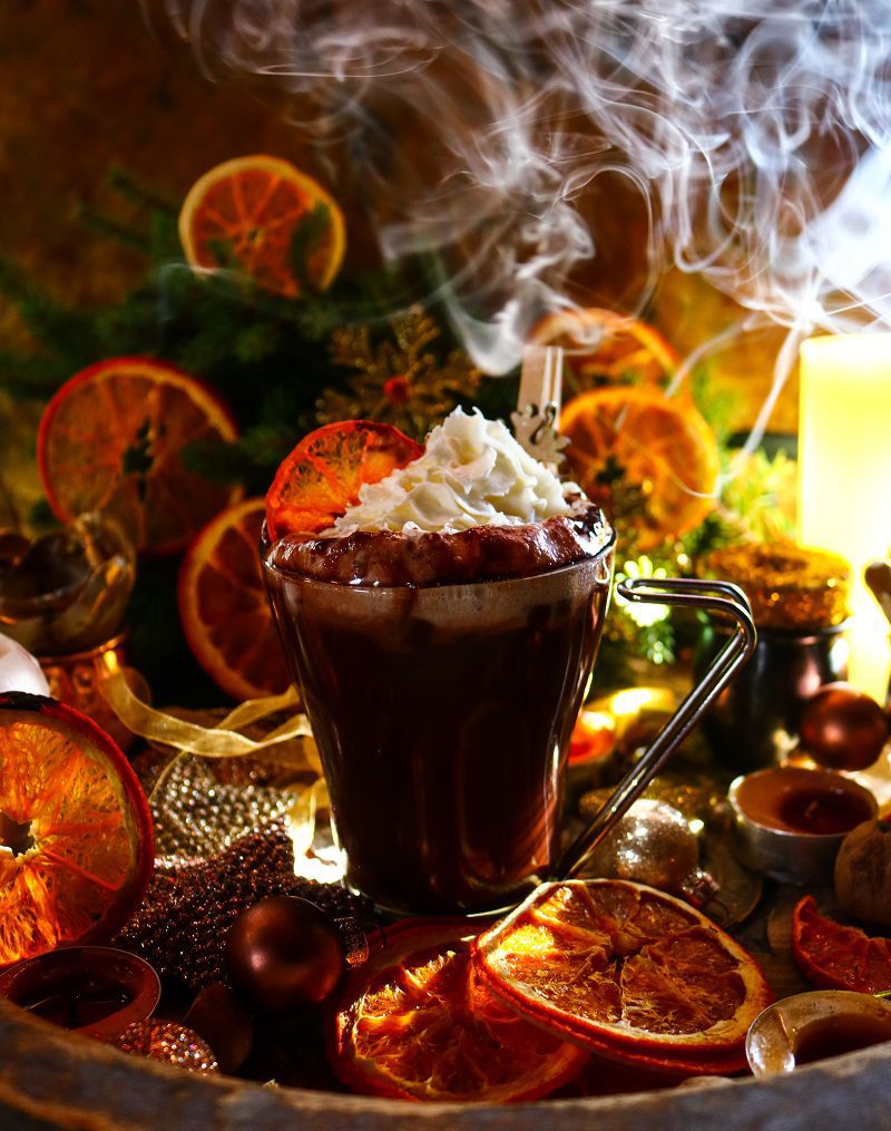 Christmas Hot Chocolate Collection: A festive cup of hot chocolate topped with whipped cream, garnished with dried orange slices and a wooden snowflake ornament, surrounded by glowing candles, sparkling decorations, and golden ornaments, creating a cozy holiday scene.