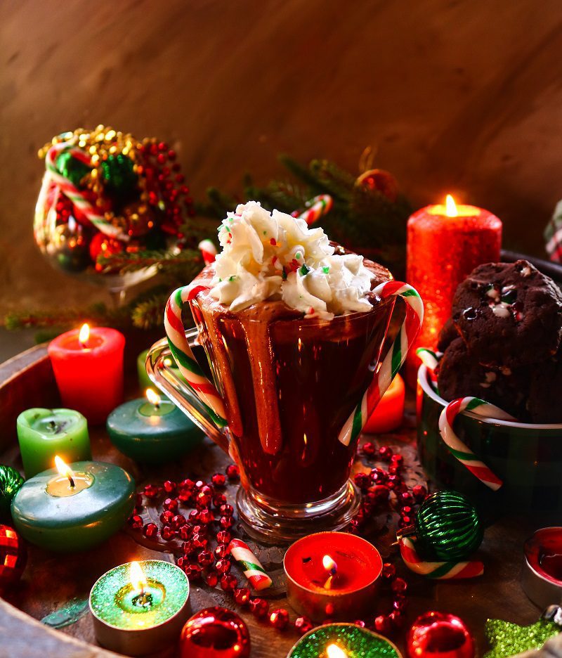 A festive holiday hot chocolate topped with whipped cream and colorful sprinkles, surrounded by glowing candles, candy canes, and chocolate cookies, creating a cozy Christmas atmosphere.