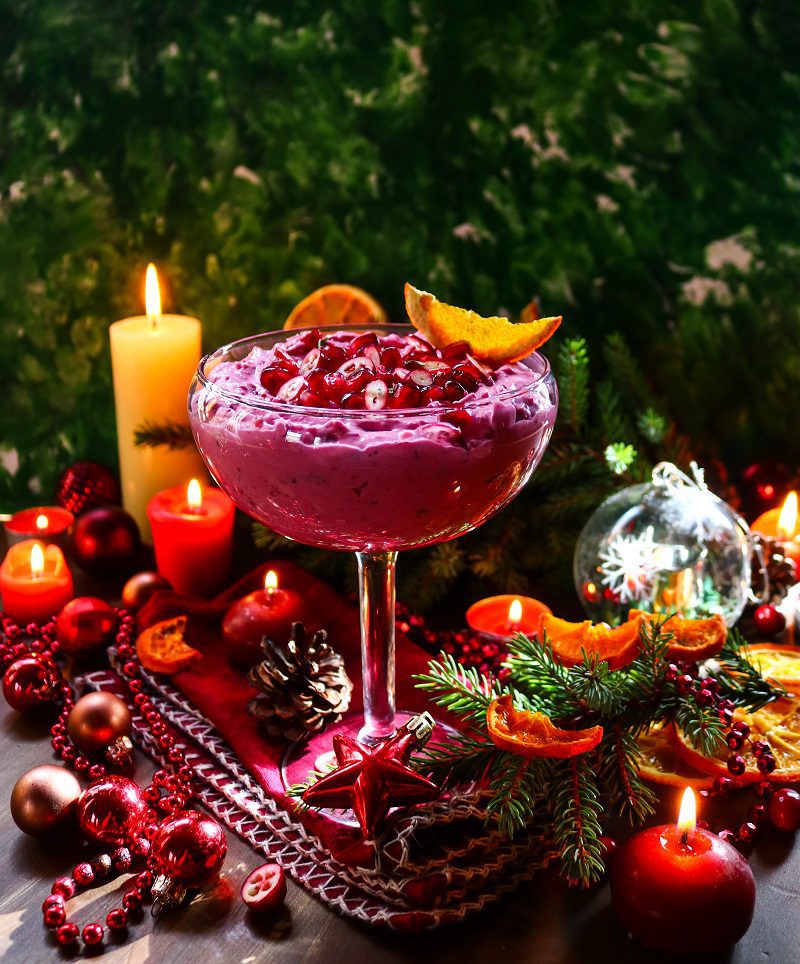 A tall glass serving dish filled with creamy cranberry goat cheese spread, garnished with cranberries and dried orange, set among glowing candles, ornaments, and evergreen branches. This holiday-inspired cranberry goat cheese spread showcases its bright colour and smooth, whipped texture, making it an elegant Christmas appetizer or festive party dip.