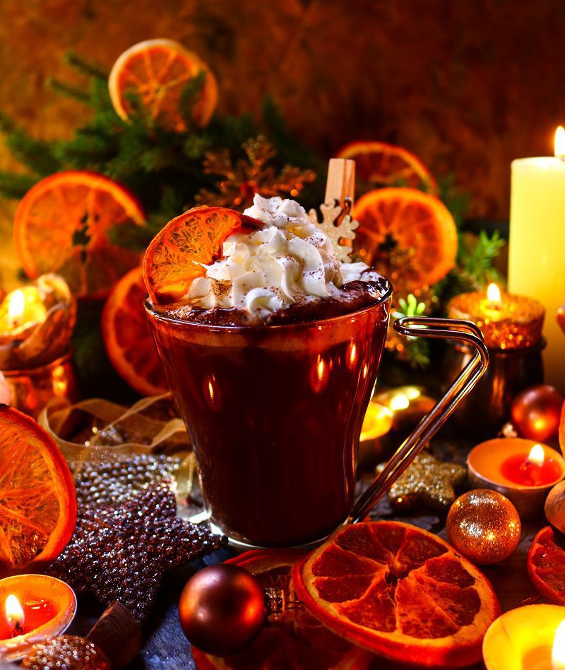 7 Festive Hot Chocolate Recipes for the Holiday Season: A cozy cup of hot chocolate topped with whipped cream, garnished with dried orange slices and a festive wooden ornament, surrounded by twinkling candles, Christmas decorations, and golden ornaments for a warm holiday ambiance.