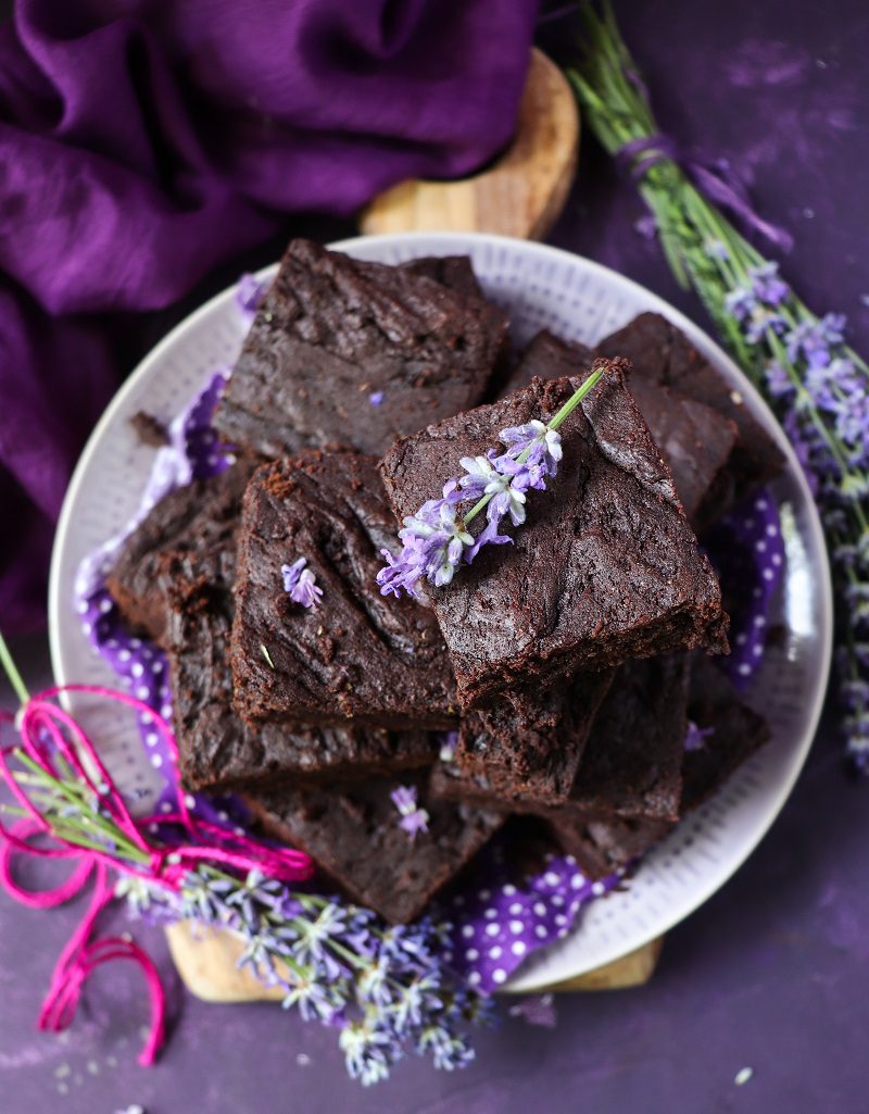 Lavender brownies stacked on a white plate, rich and fudgy dark chocolate squares garnished with fresh lavender sprigs on a purple backdrop.