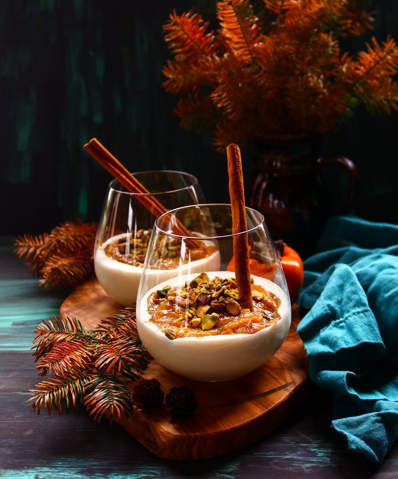 No bake persimmon dessert with a creamy cheesecake-style base, persimmon topping, pistachios, and cinnamon sticks served in glass cups.