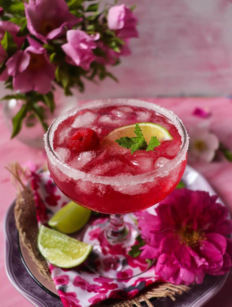 Chilled rose vermouth cocktail with crushed ice and lime in a sugar-rimmed glass, served on a tray with fresh limes and pink flowers.