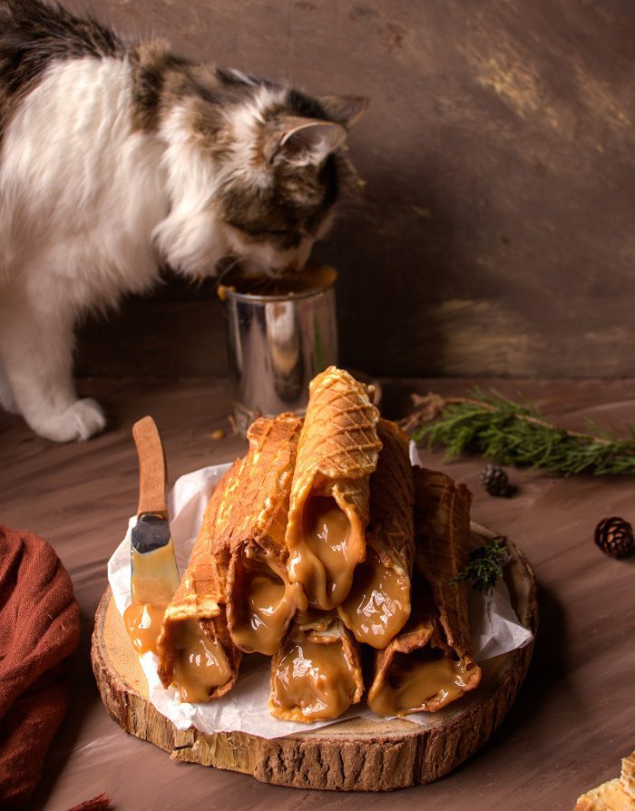 Rolled waffle cookies filled with dulce de leche on a wooden board, with a curious cat in the background.