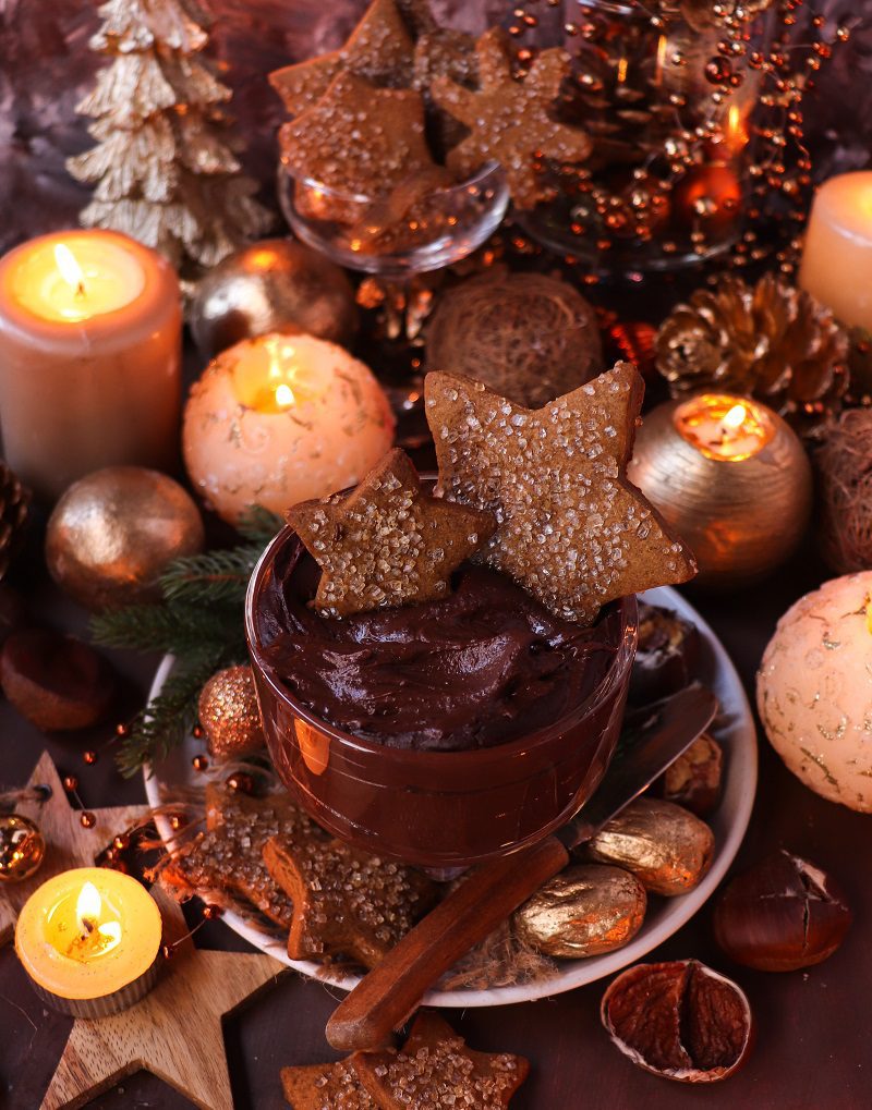 A festive glass dish filled with rich chocolate chestnut dessert spread, garnished with sparkling gingerbread star cookies and surrounded by glowing candles, gold ornaments, and Christmas decorations for a warm holiday atmosphere.