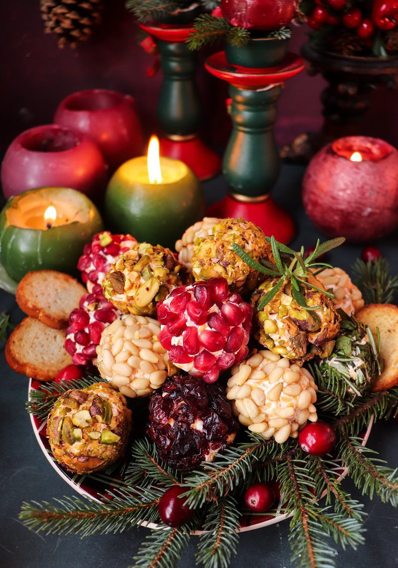 A festive platter of mini cheese balls appetizer coated in pistachios, cranberries, pine nuts, and pomegranate arils, surrounded by evergreen sprigs and cranberries — perfect for Christmas entertaining.