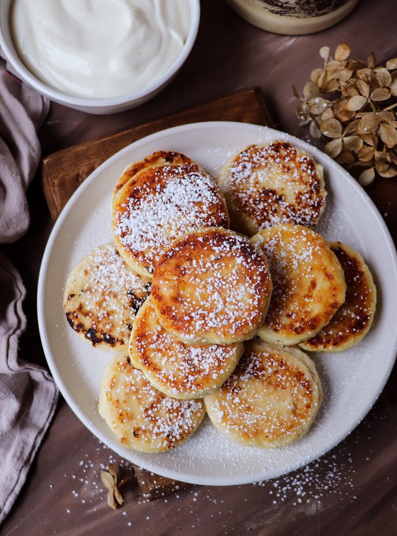 Overhead view of golden pan-fried cottage cheese pancakes arranged on a white plate, dusted with icing sugar, with a bowl of sour cream nearby.