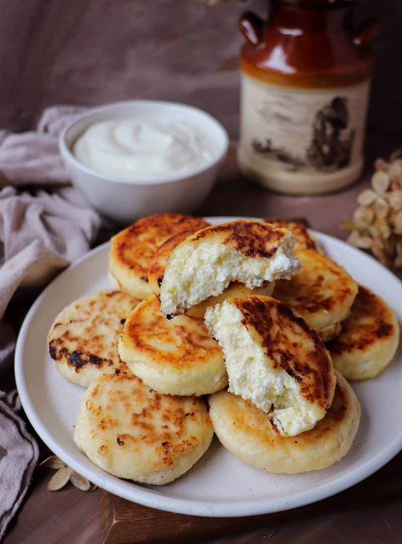 Pan-fried cottage cheese pancakes stacked on a white plate, with one pancake split open to show the soft, creamy interior, served with a bowl of sour cream in the background.