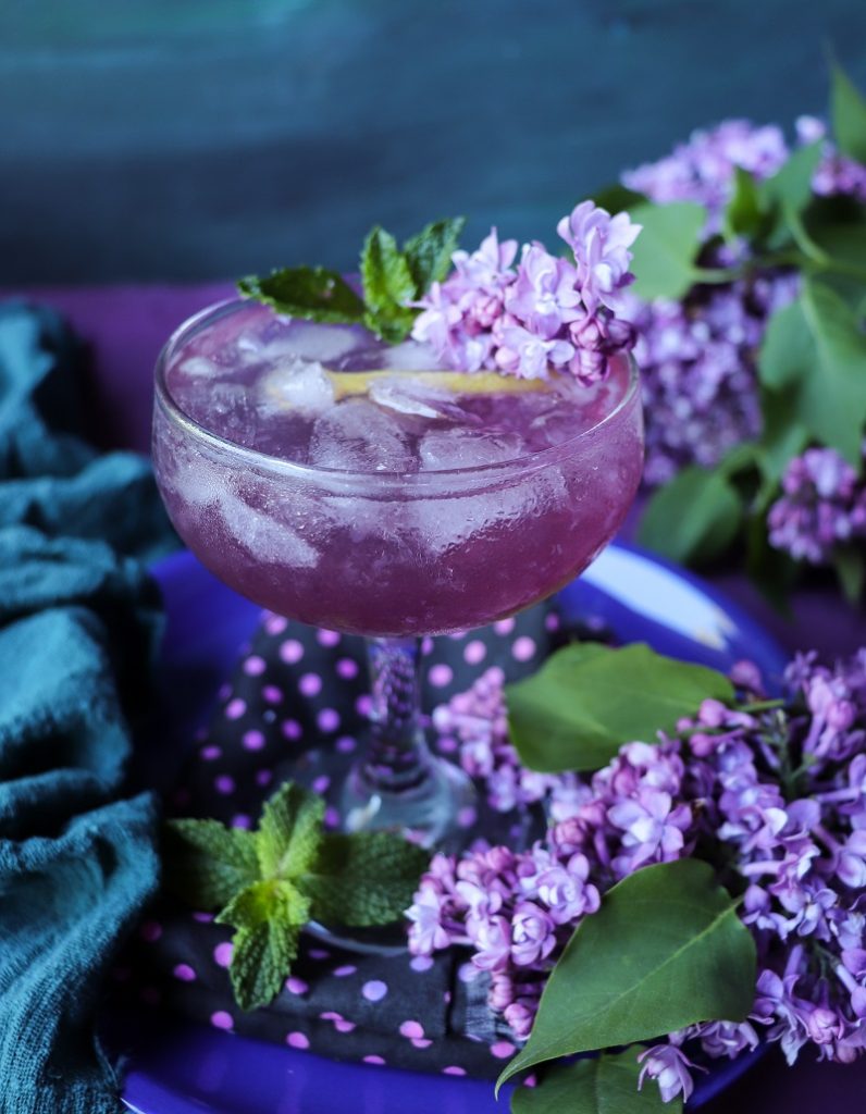 Lilac Empress gin cocktail with lilac syrup, lemon, and mint served in a coupe glass, surrounded by fresh lilac blossoms.