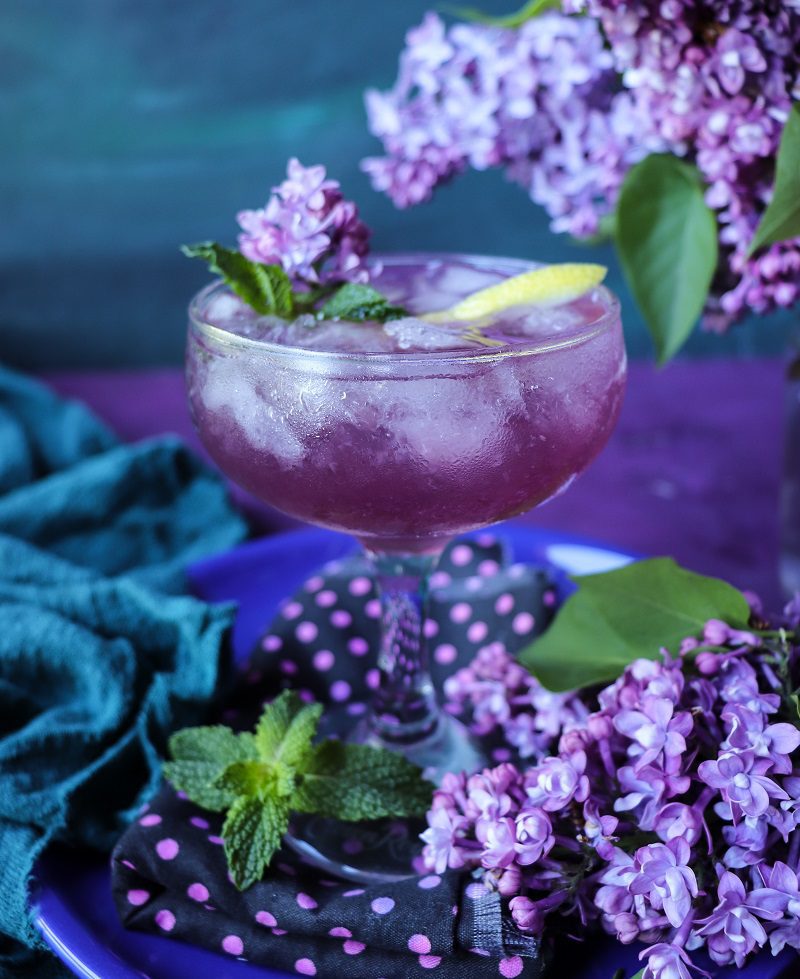 Lilac Empress gin cocktail with lilac syrup, lemon, and mint served in a coupe glass, surrounded by fresh lilac blossoms.