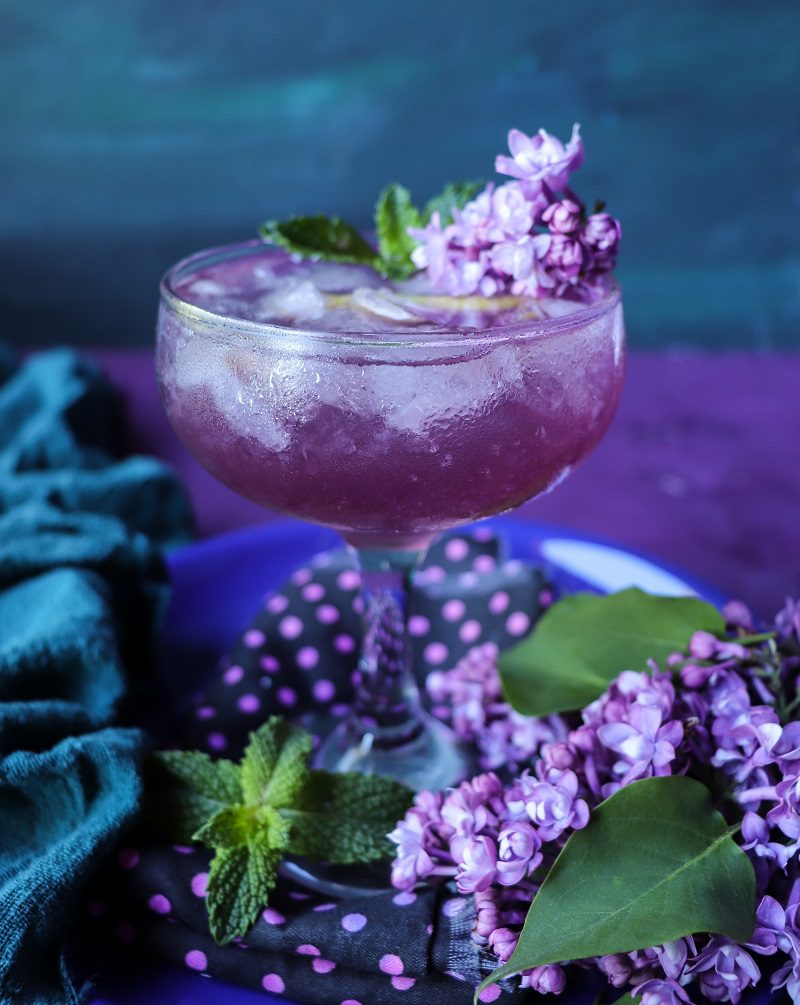 Lilac Empress gin cocktail with lilac syrup, lemon, and mint served in a coupe glass, surrounded by fresh lilac blossoms.