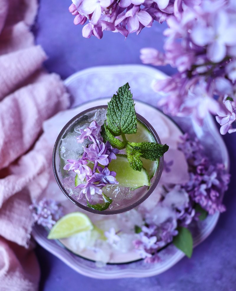 Lilac mojito served in a tall glass with crushed ice, mint leaves, and lilac blossoms, showing a soft purple-pink gradient drink, garnished with fresh lilac flowers and a lime wedge on a floral-styled plate.