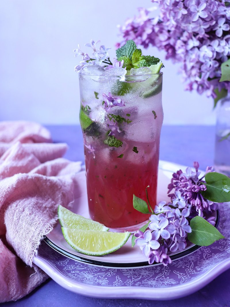 Lilac mojito served in a tall glass with crushed ice, mint leaves, and lilac blossoms, showing a soft purple-pink gradient drink, garnished with fresh lilac flowers and a lime wedge on a floral-styled plate.