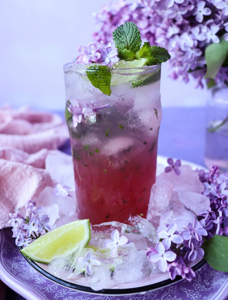 Lilac mojito served in a tall glass with crushed ice, mint leaves, and lilac blossoms, showing a soft purple-pink gradient drink, garnished with fresh lilac flowers and a lime wedge on a floral-styled plate.