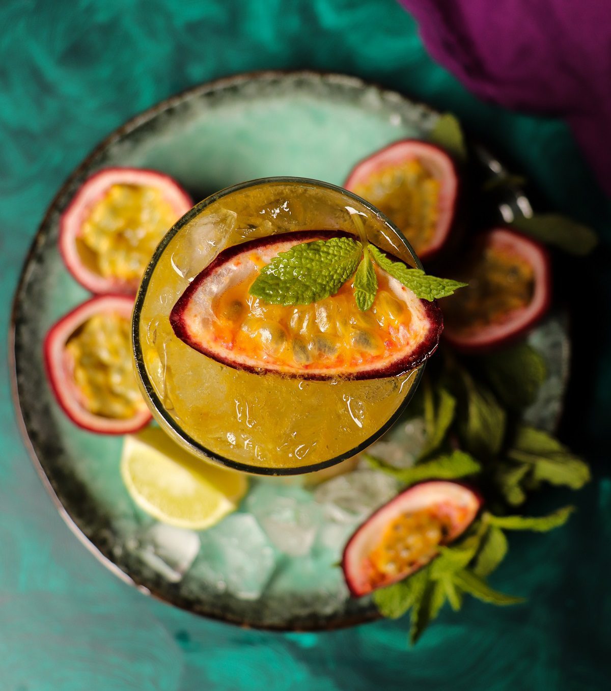 Overhead view of a passion fruit mojito cocktail with crushed ice, bright yellow passion fruit pulp, mint garnish, and a sliced passion fruit on top, styled with lime wedges and mint on a teal surface.