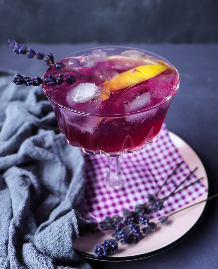Lavender Empress 1908 gin cocktail in a coupe glass, filled with ice and showing a deep purple-pink hue, garnished with a lemon slice and fresh lavender sprigs, styled on a soft pink plate with a purple gingham napkin and dried lavender.