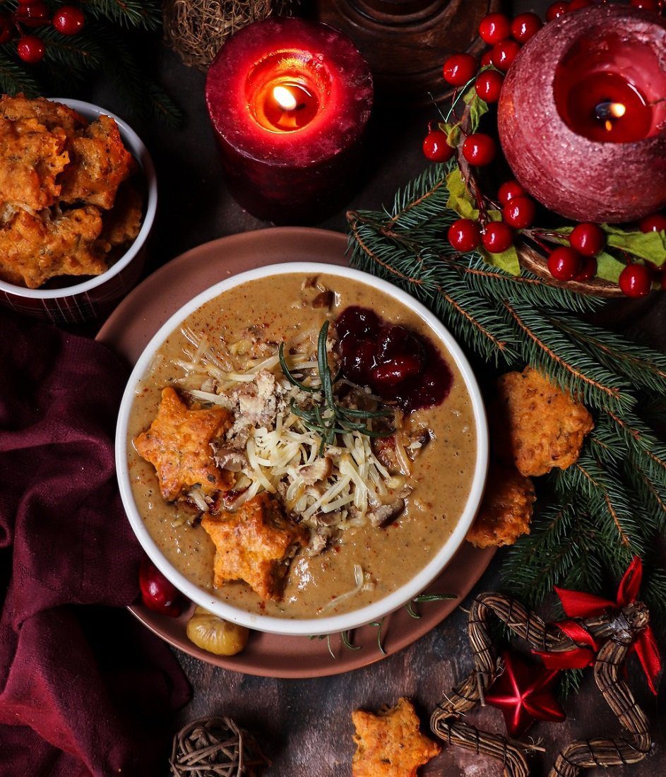 Overhead view of Creamy Chestnut Soup with rosemary and smoked cheddar, topped with shredded cheese, cranberry sauce, and cheese biscuits, surrounded by pine branches and cranberries — a cozy and elegant creamy chestnut cheese soup perfect for winter entertaining.