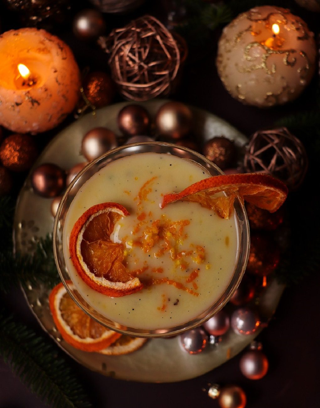 A creamy white chocolate martini made with real melted white chocolate, topped with fresh orange zest and garnished with two dried orange slices. The drink sits against a festive backdrop of holiday ornaments and warm candlelit tones, creating a cozy Christmas atmosphere.