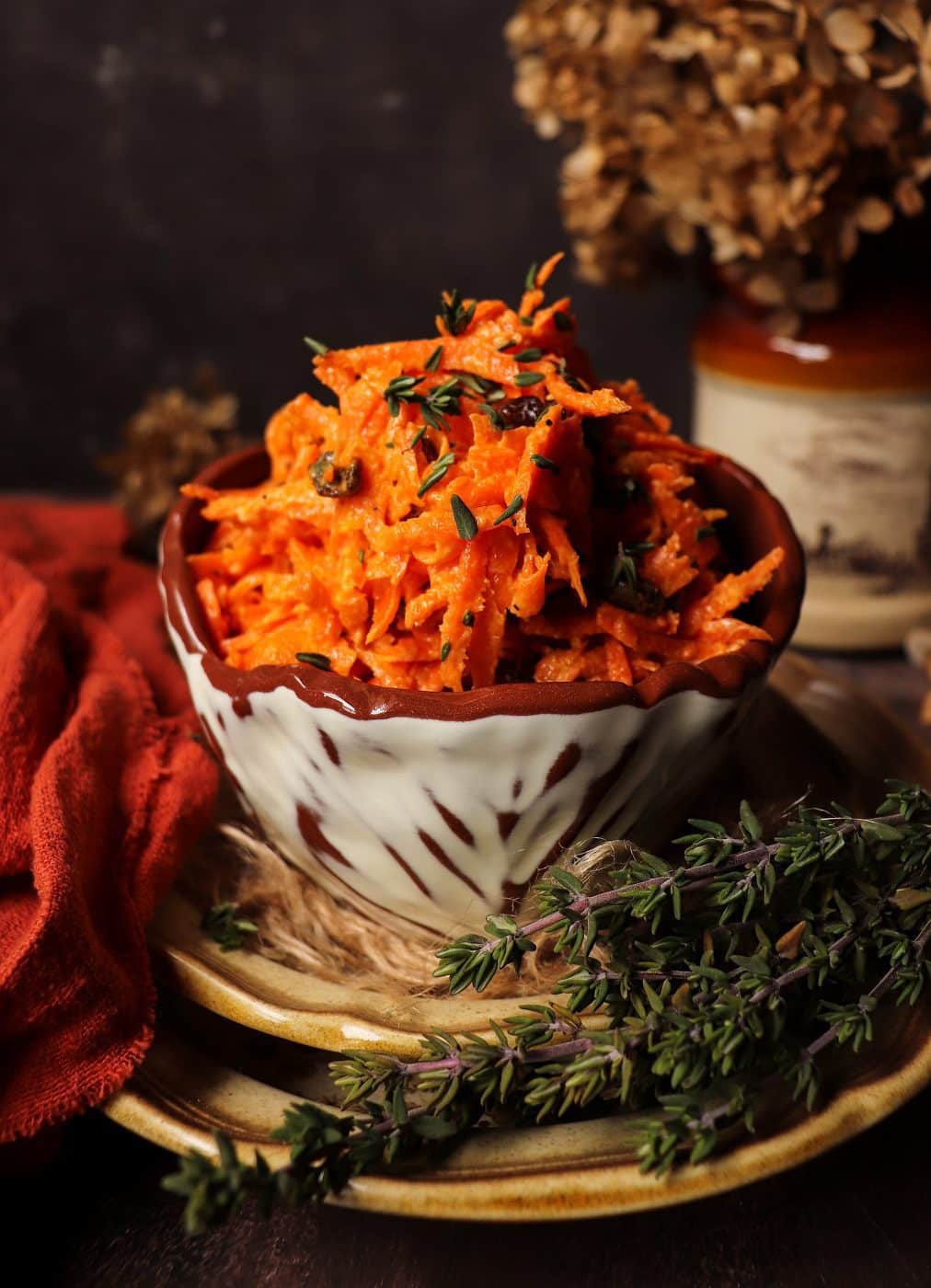 Carrot raisin salad served in a rustic ceramic bowl, topped with fresh thyme leaves and surrounded by thyme sprigs on a wooden surface with a warm, moody background.