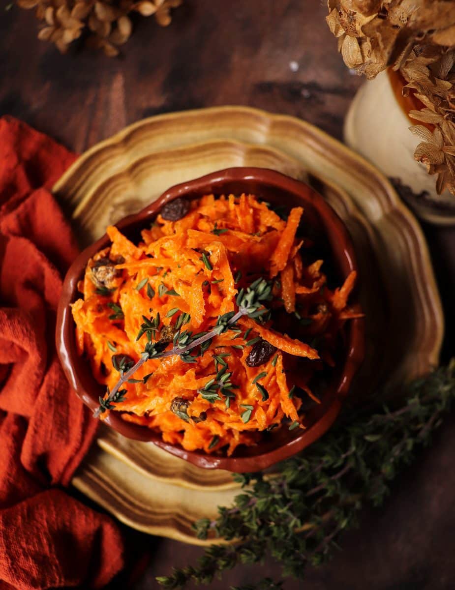 Carrot raisin salad served in a rustic ceramic bowl, topped with fresh thyme leaves and surrounded by thyme sprigs on a wooden surface with a warm, moody background.