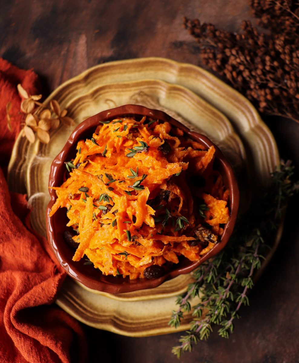 Carrot raisin salad served in a rustic ceramic bowl, topped with fresh thyme leaves and surrounded by thyme sprigs on a wooden surface with a warm, moody background.