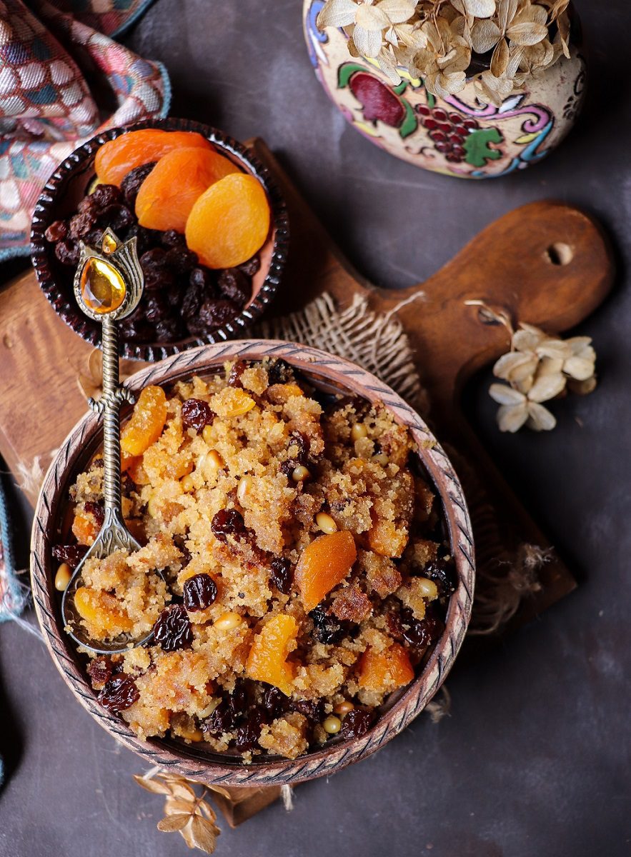 Semolina halwa (sooji halwa) with raisins, dried apricots, and pine nuts served in a decorative bowl with a spoon.