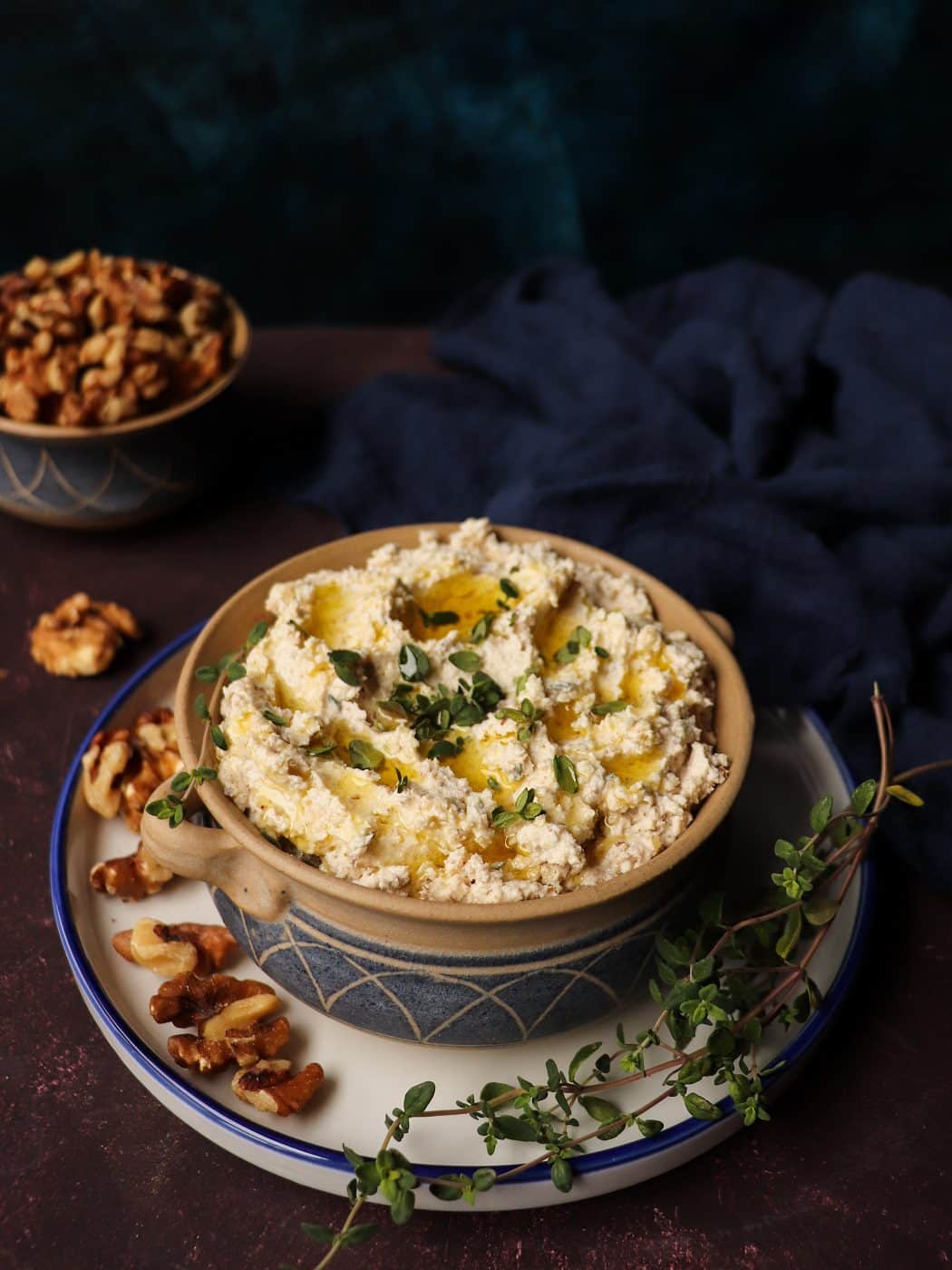 Walnut feta dip topped with toasted walnut halves and fresh thyme, served in a rustic ceramic bowl with olive oil drizzle, surrounded by extra walnuts and bread rolls in the background.