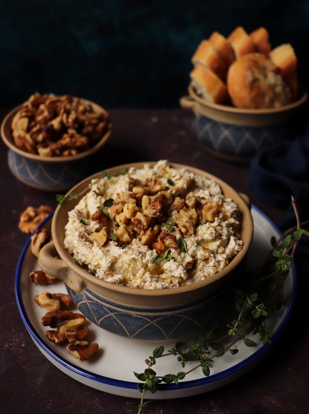 Walnut feta dip topped with toasted walnut halves and fresh thyme, served in a rustic ceramic bowl with olive oil drizzle, surrounded by extra walnuts and bread rolls in the background.