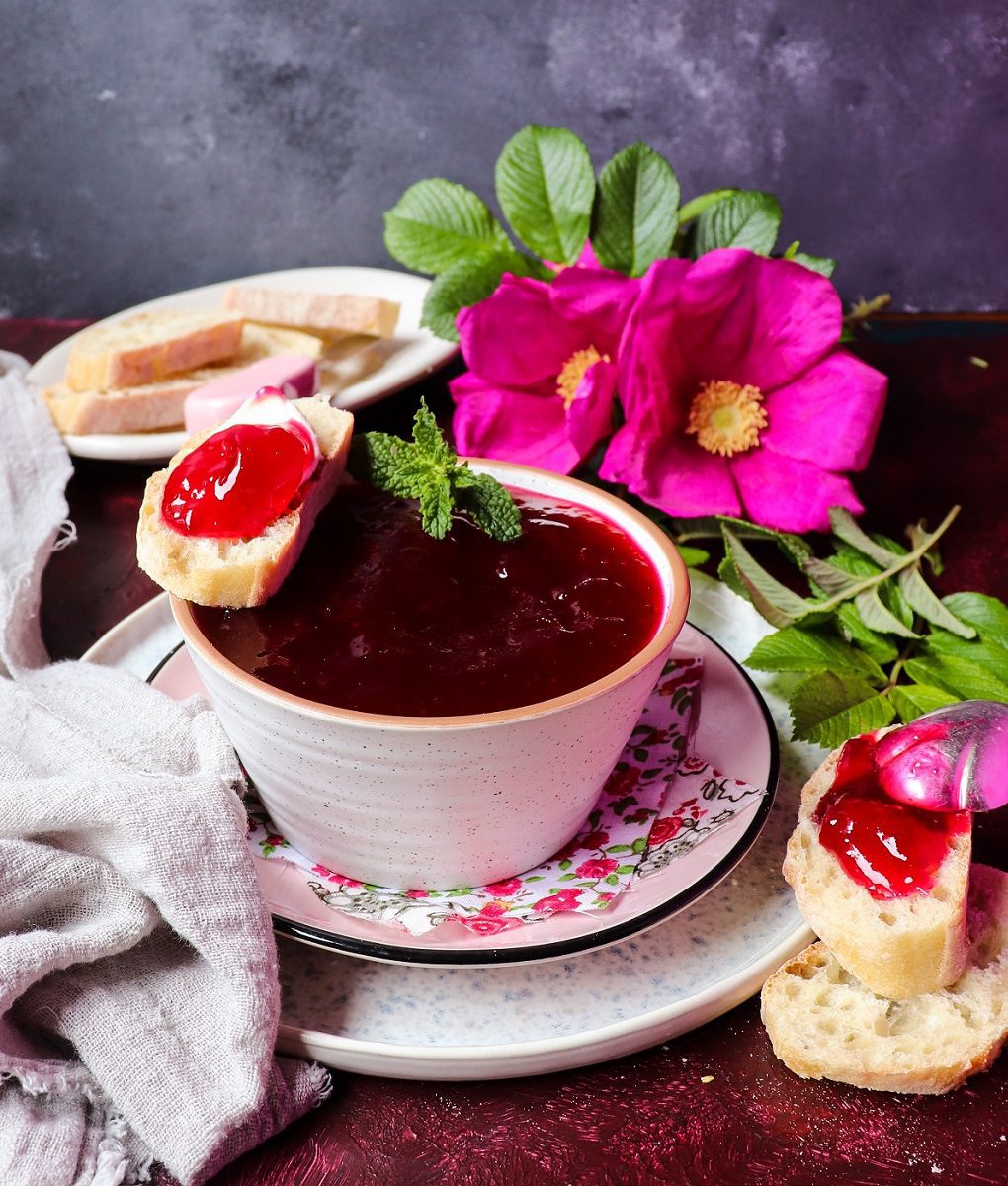 Bright red rose petal jelly in a bowl with a spoon, served with crusty bread and fresh wild rose blossoms.