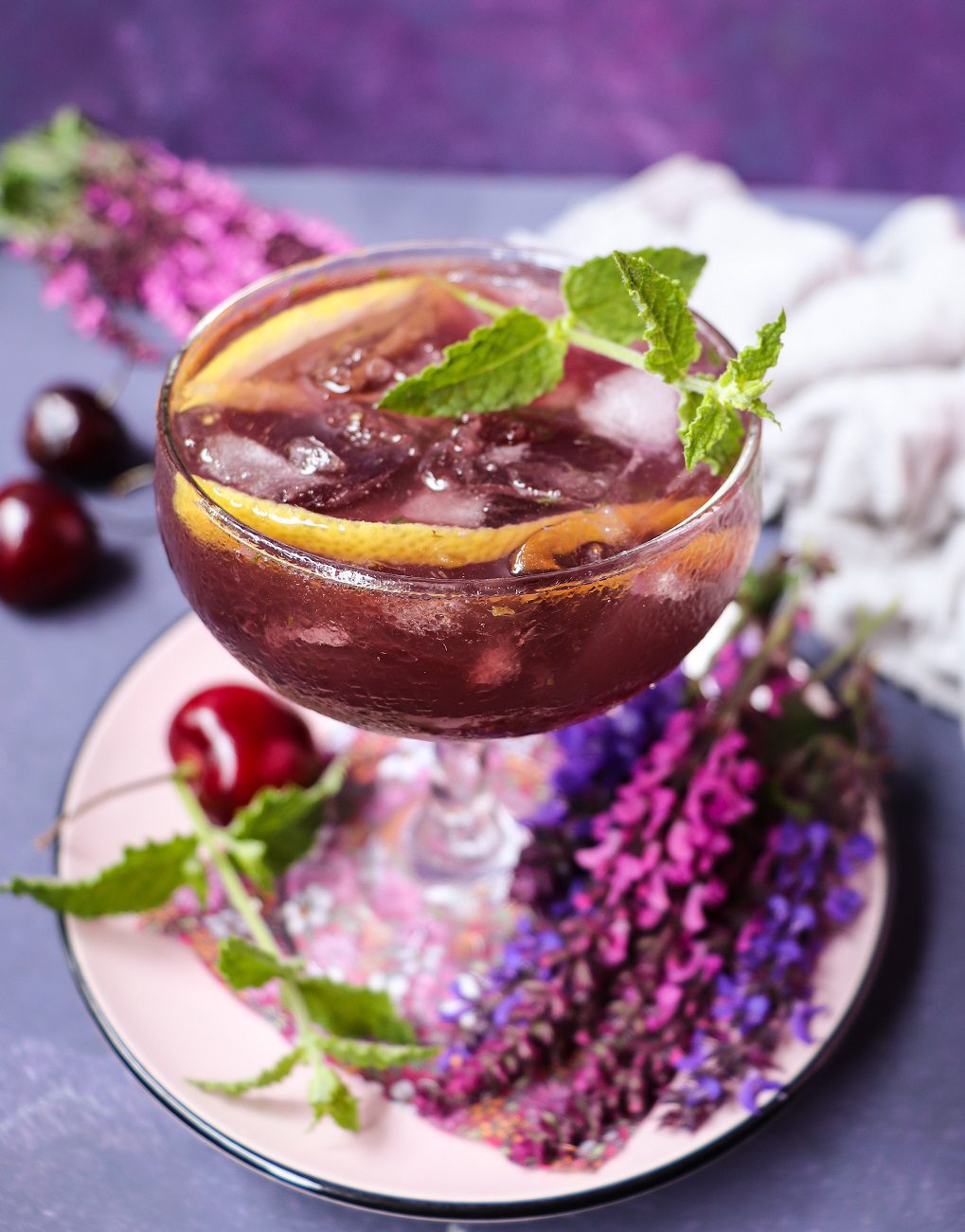 Cherry gin cocktail with Empress 1908 Gin, served over ice in a coupe glass and garnished with fresh mint, cherries, and citrus peel.